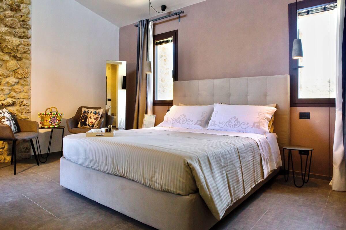 A bedroom with a large bed, a small table, a chair, and a stone wall.