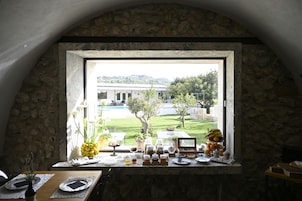 Un comedor con pared de piedra, una ventana con vista a un edificio y un jardín, y una mesa dispuesta con platos, cubiertos y una bandeja de fruta.