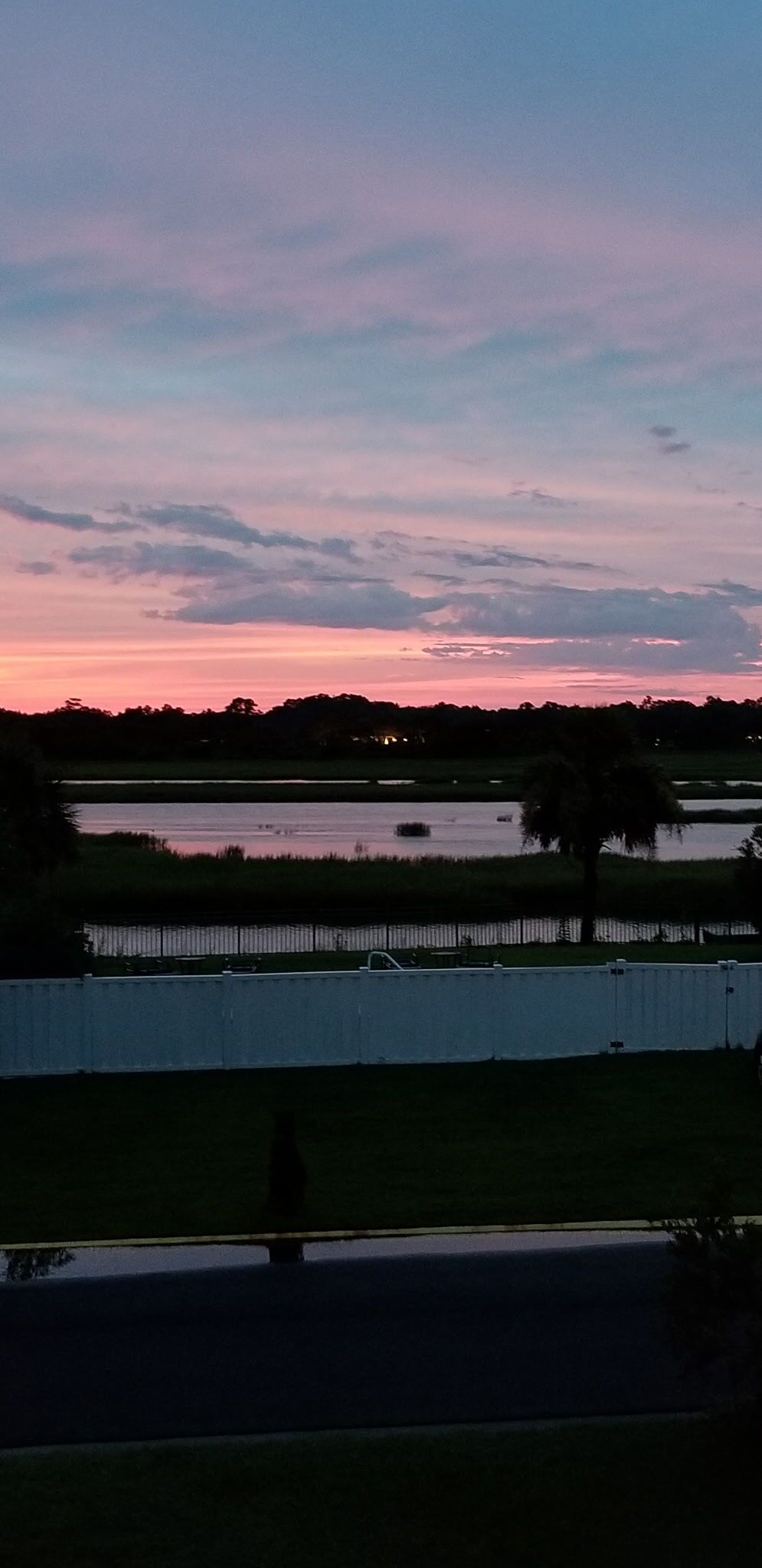 A sunset over a body of water with a fence and a solitary figure in the foreground.
