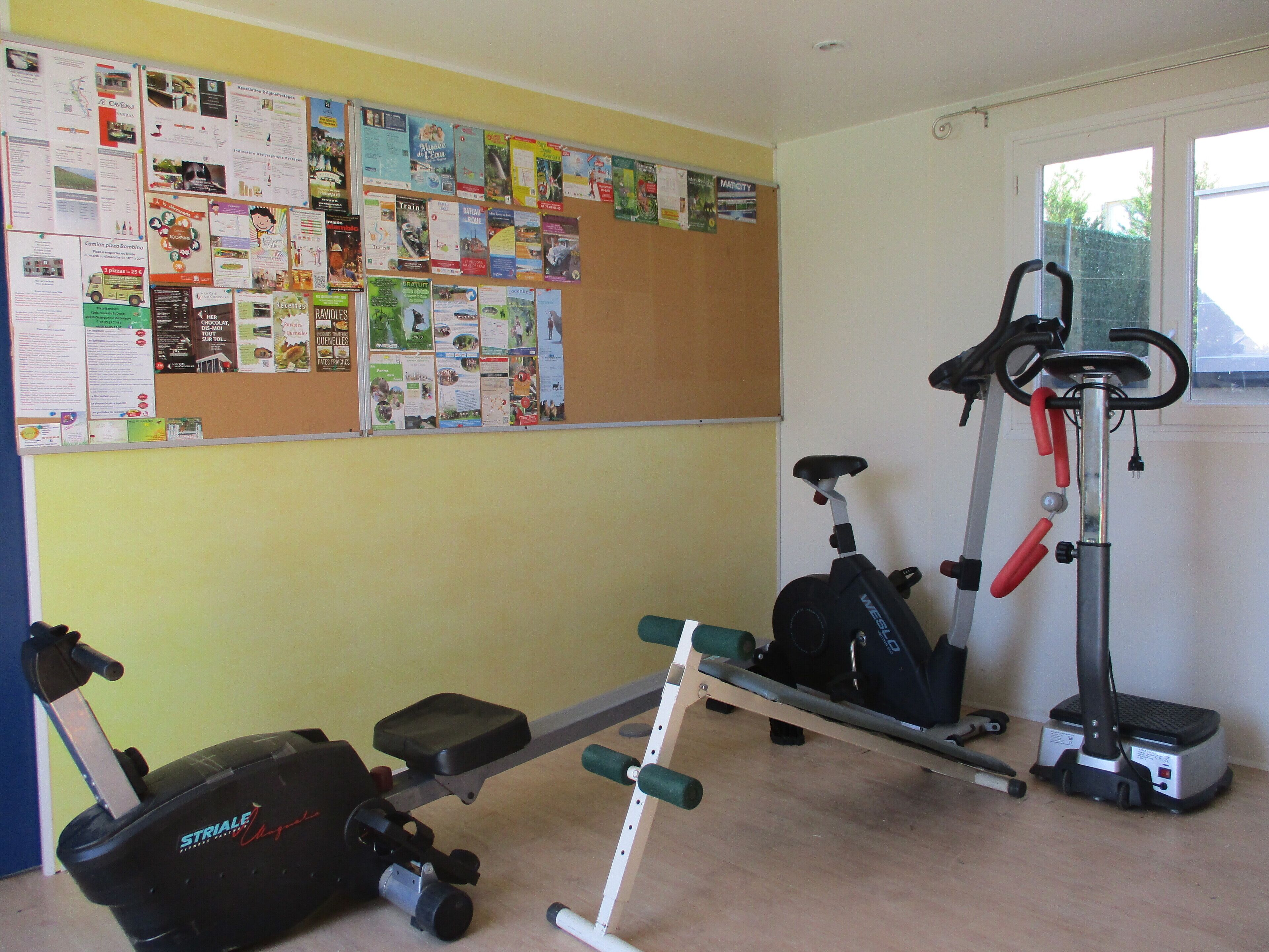 Un gimnasio en casa con equipo de ejercicio, un tablón de anuncios con varios avisos y una ventana con vistas al exterior.