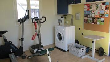 A compact home gym with a stationary bike, a treadmill, a washing machine, and a small workout area with a bench and weights.