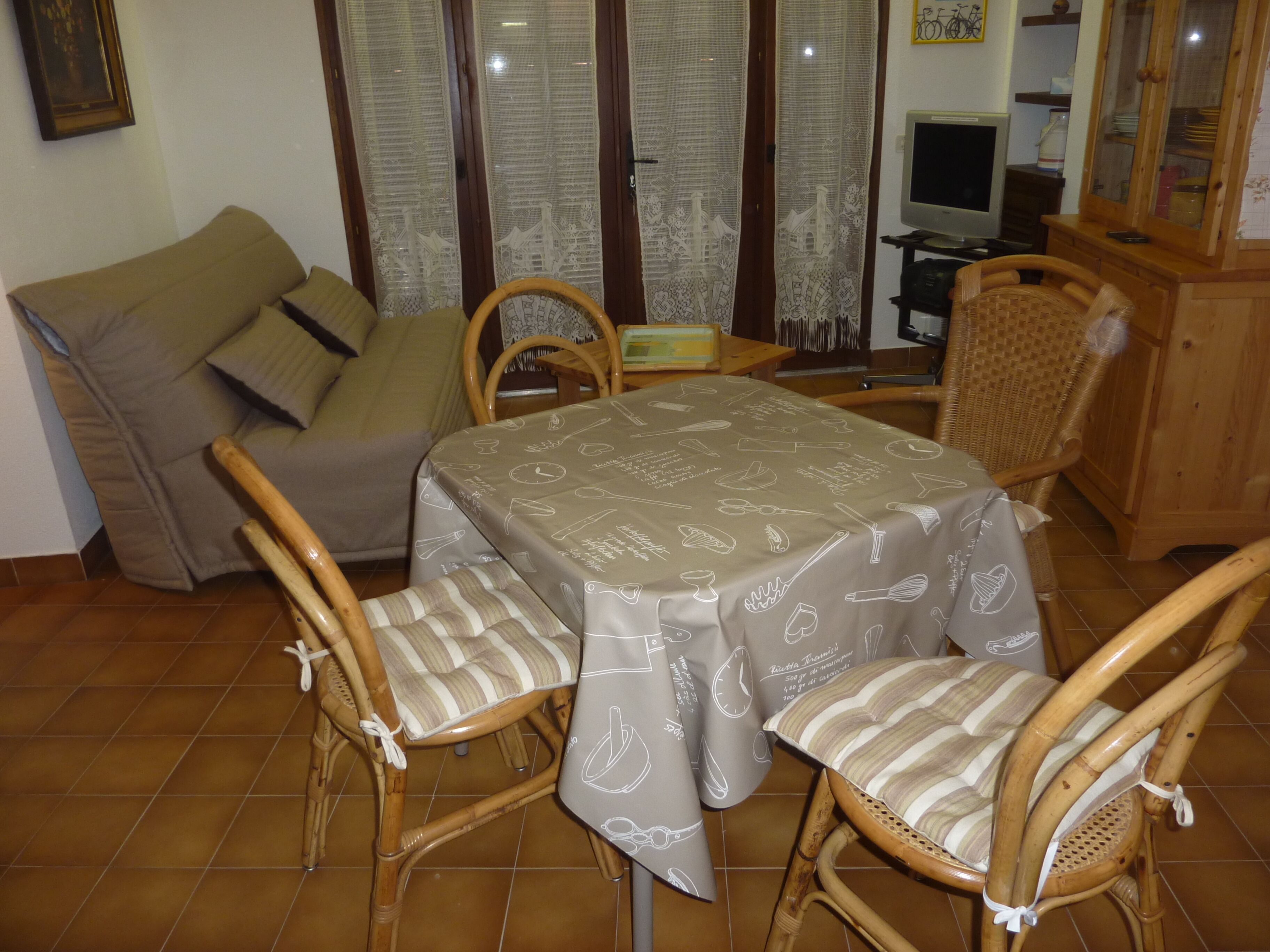 Uma sala de jantar com uma mesa coberta por toalha, utensílios de cozinha e cadeiras de vime com almofadas listradas.