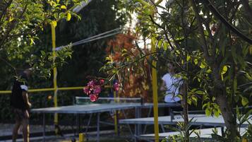 Een tennisbaan met tafels en stoelen, omringd door bomen en een gele hekken.