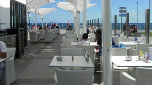 An outdoor dining area with white umbrellas, tables, and chairs, overlooking the ocean. - Meloneras Beach Hh1010 Maspalomas Duplex (San Bartolomé de Tirajana)