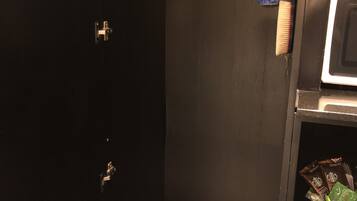 Pequeña habitación oscura con una puerta negra, un perchero metálico con ganchos, un atomizador y una cesta con toallas blancas.