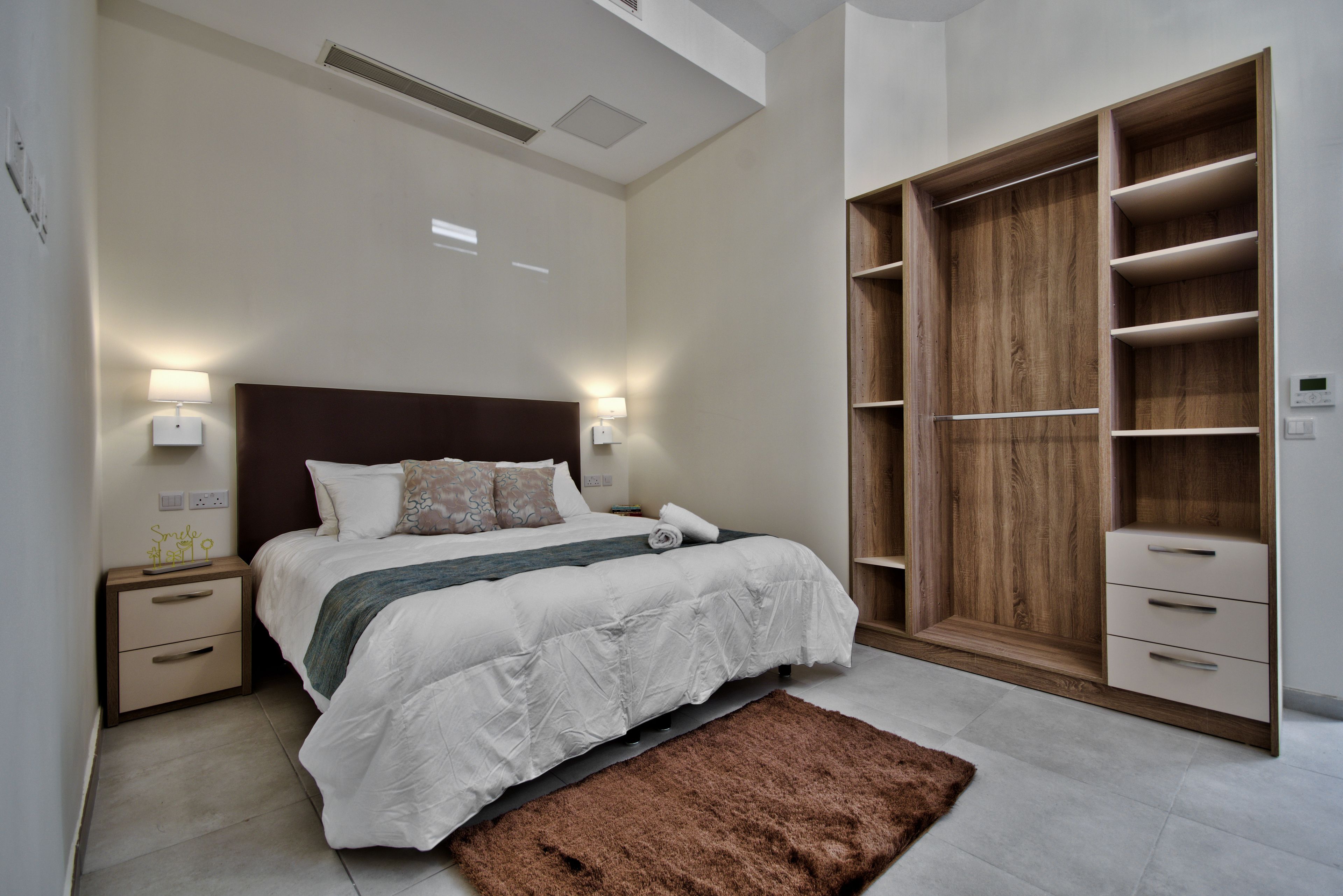 Un dormitorio con una cama grande, lámparas de noche, un armario de madera y un tapete marrón.