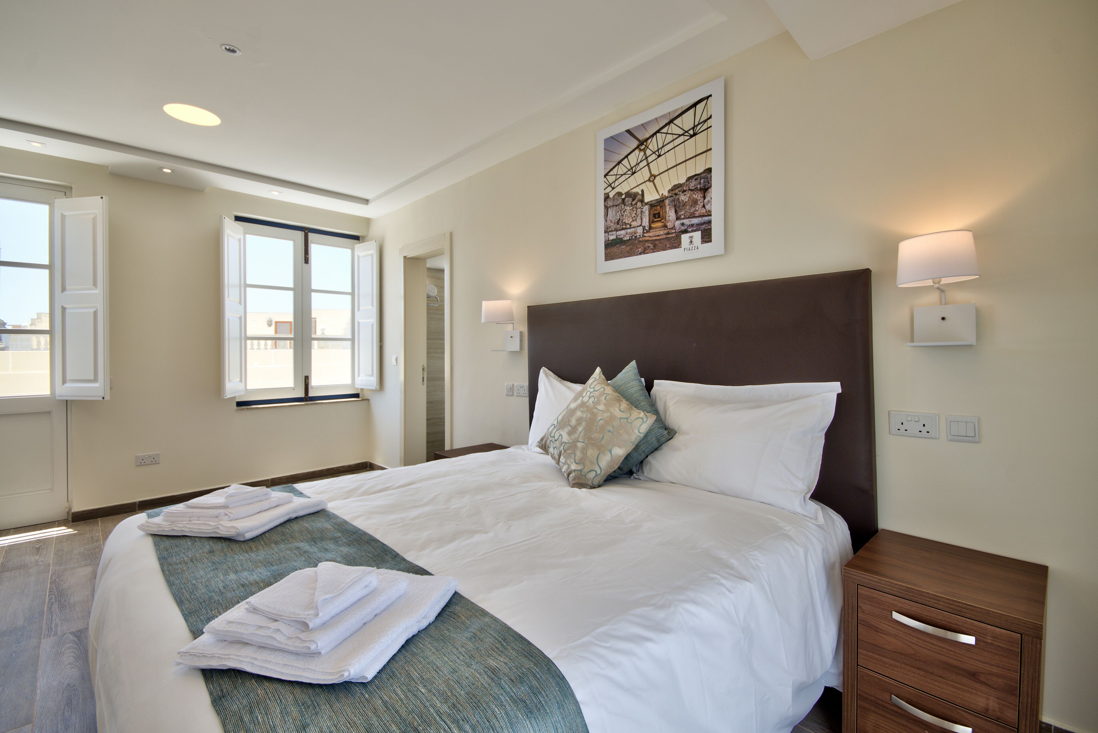 A hotel room with a large bed, a headboard, bedside lamps, a window, and a painting on the wall.