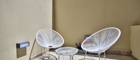 Una pequeña zona de estar al aire libre con dos sillas blancas de estructura metálica y una mesa pequeña, frente a una pared color crema con una maceta de flores.