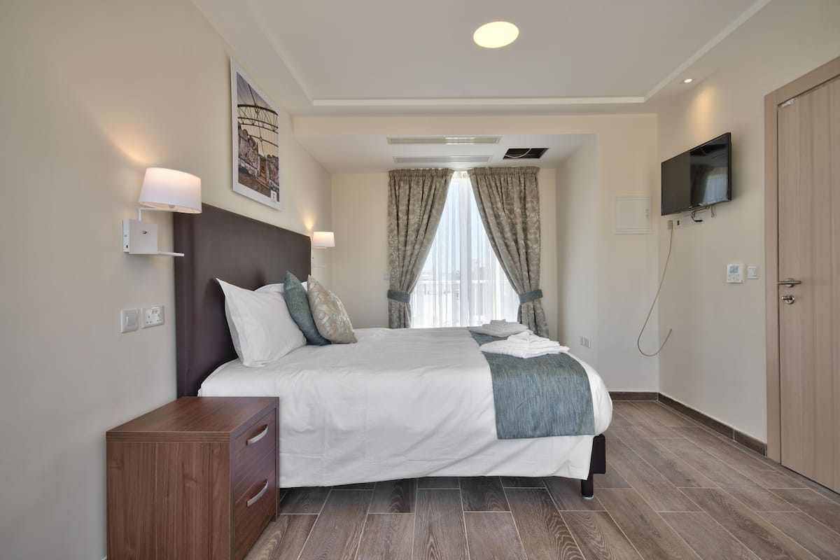 A modern hotel room with a large bed, a wooden nightstand, a flat-screen TV, and a window with curtains.