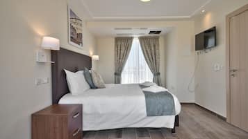 Una habitación de hotel moderna con una cama grande, una mesita de noche de madera, un televisor de pantalla plana y una ventana con cortinas.