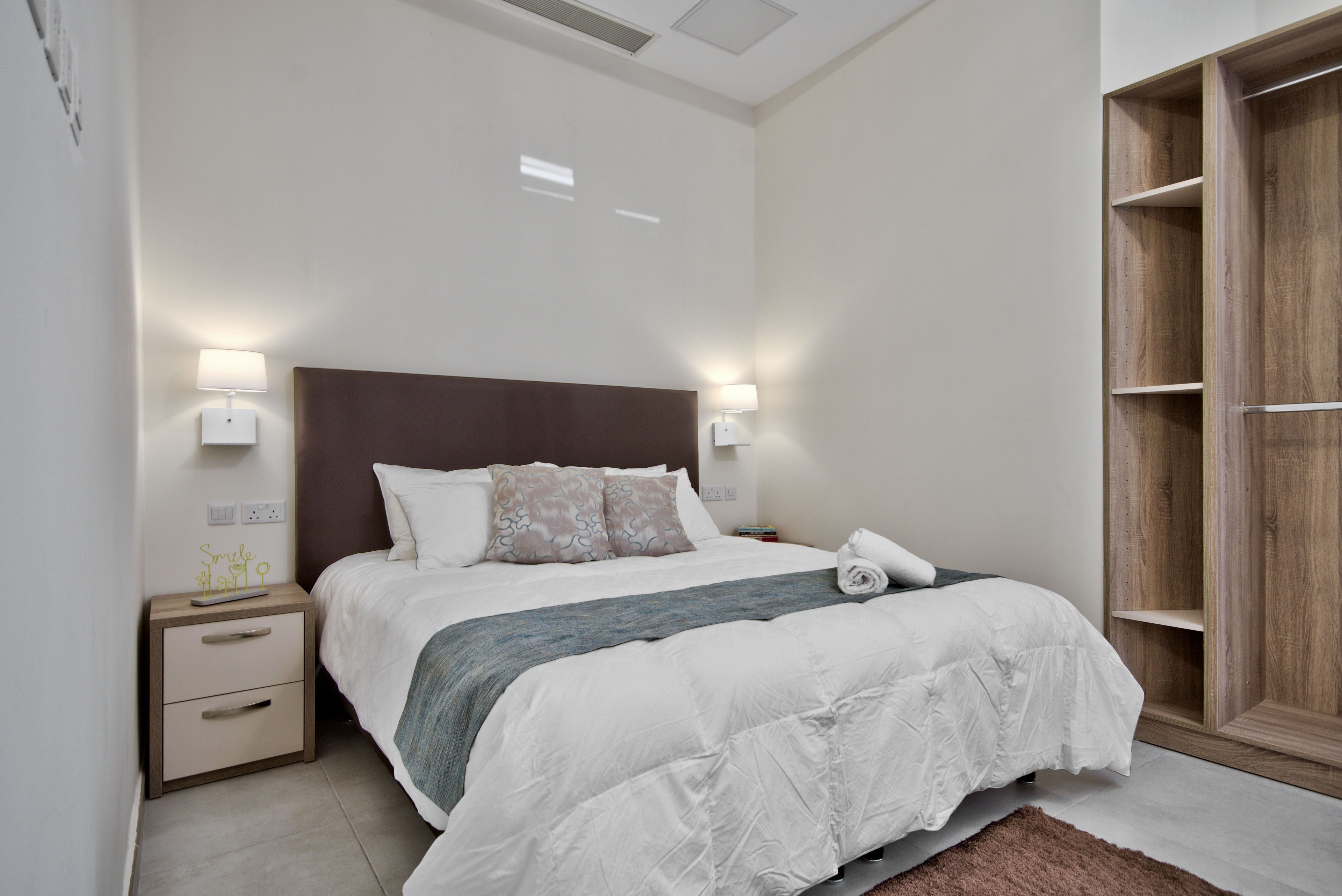 A neatly made bed with a wooden headboard, bedside lamps, a nightstand, and a wooden wardrobe.
