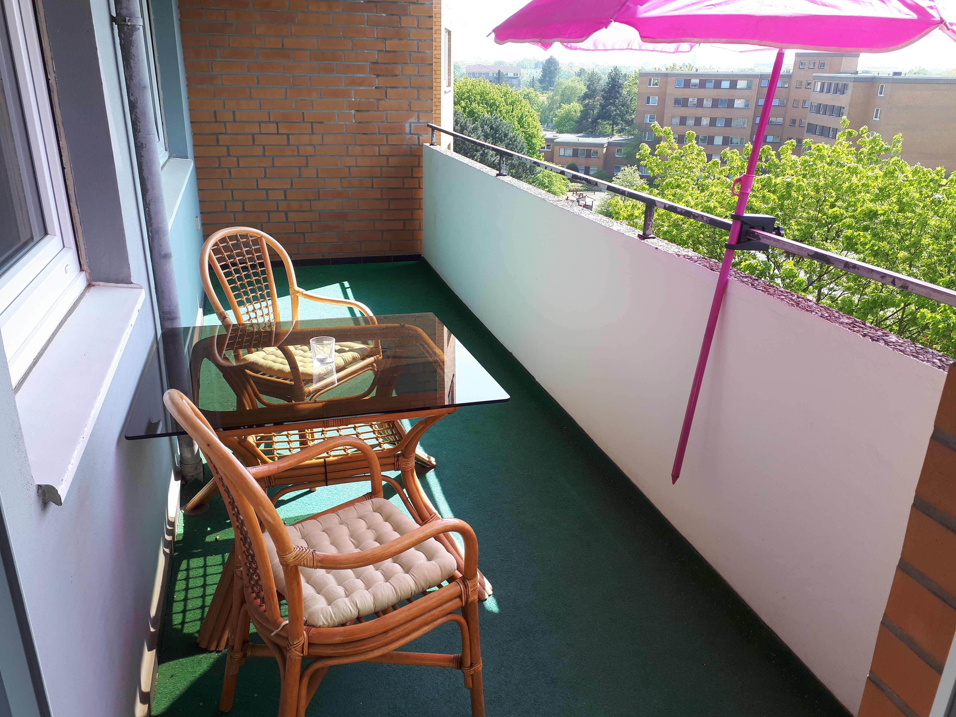 A balcony with a glass table and wicker chairs, a pink umbrella, and a view of buildings and trees.