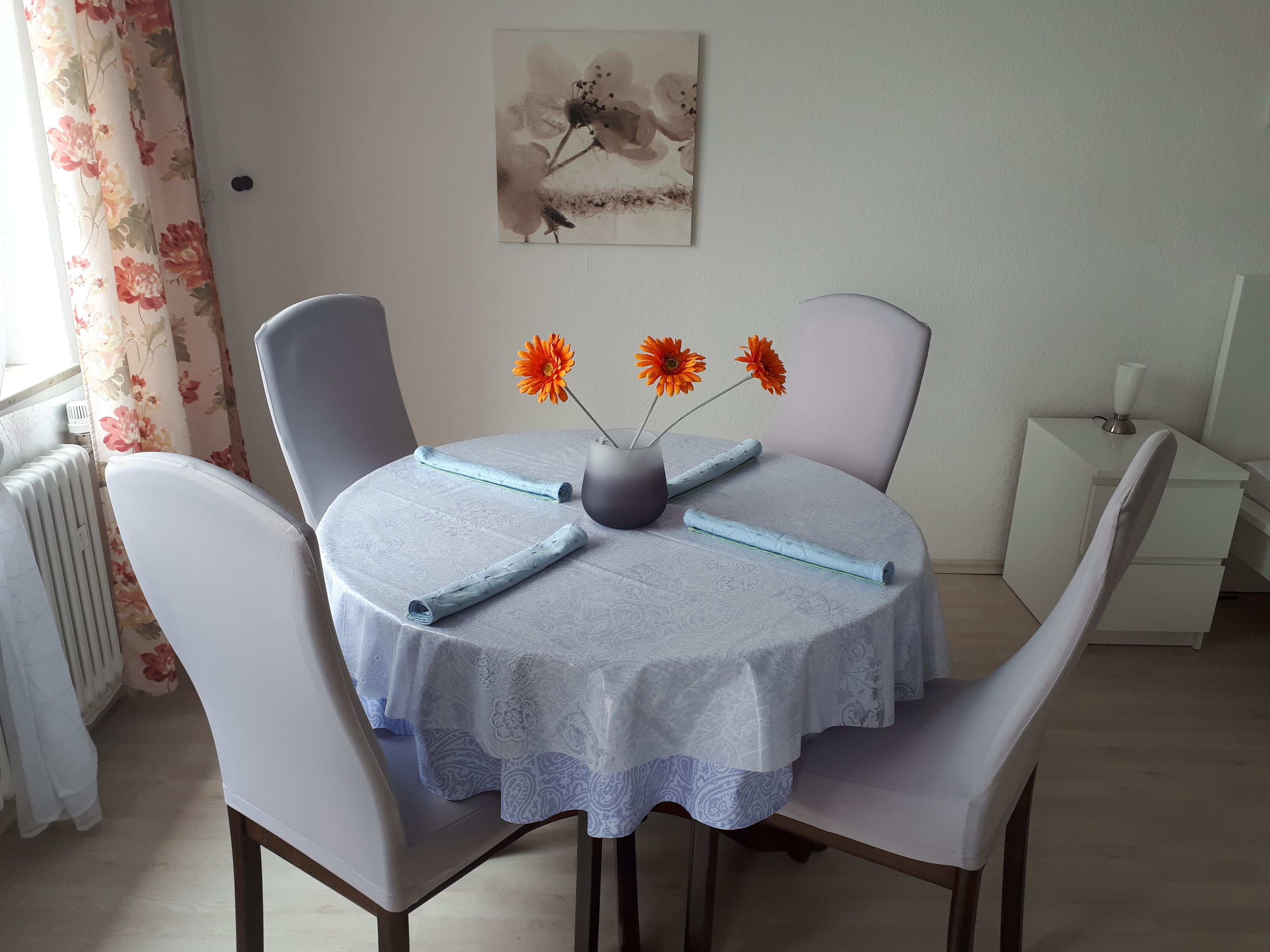 A round dining table with a white lace tablecloth, two white chairs, and a vase with orange flowers.