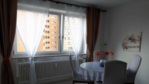 Un coin repas avec une table et des chaises, une fenêtre avec des rideaux, un radiateur et un tableau accroché au mur.