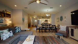 A spacious living room with a gray sofa, a coffee table, and a dining area with a wooden table and chairs. A kitchen with white cabinets and stainless steel appliances is visible in the background. - Shared Heated Pool with Water Slide, Golf Cart Access (Port Aransas)