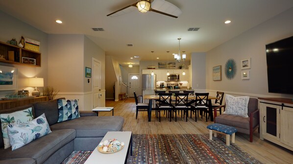 A spacious living room with a gray sofa, a coffee table, and a dining area with a wooden table and chairs. A kitchen with white cabinets and stainless steel appliances is visible in the background. - Shared Heated Pool with Water Slide, Golf Cart Access (Port Aransas)