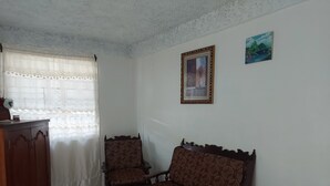 A room with a wooden cabinet, two patterned chairs, a small table, and a window with a curtain.