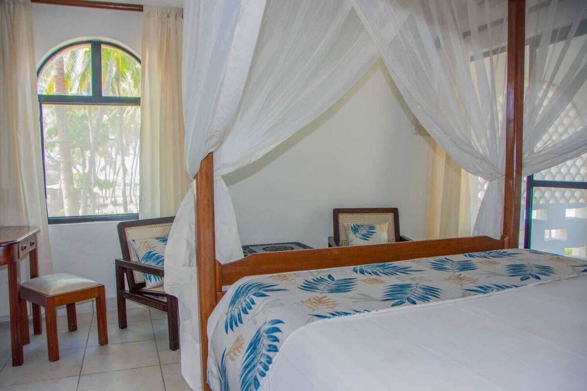 Une chambre à coucher avec un lit à baldaquin, une chaise, une petite table et une fenêtre avec des rideaux.