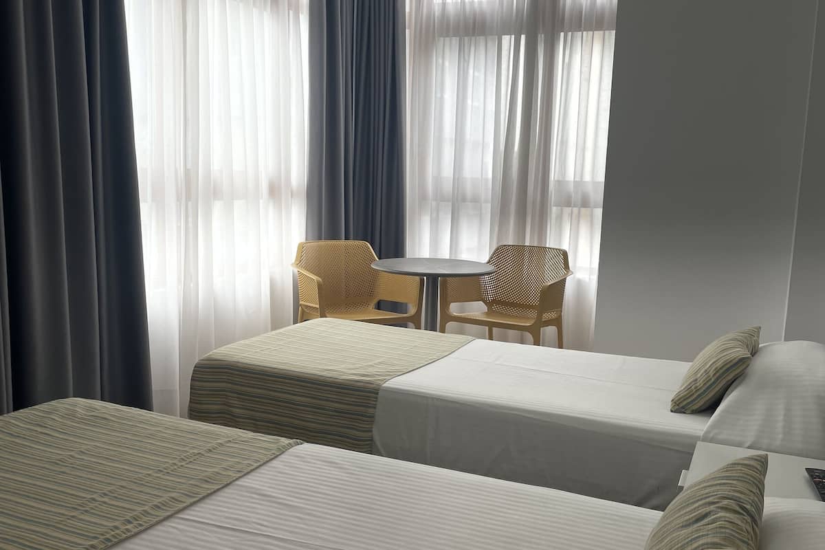 A hotel room with two single beds, a small table with two chairs, and a large window with sheer curtains.