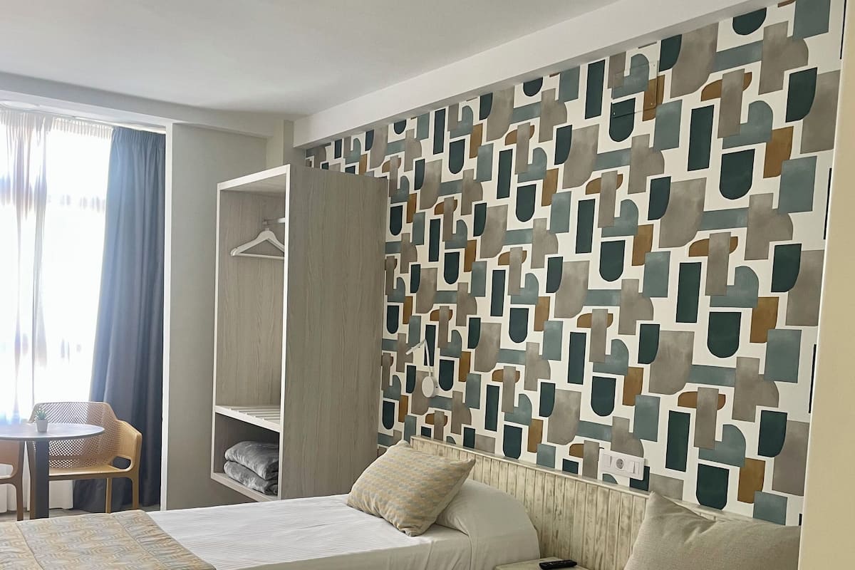 A hotel room with two beds, a wardrobe, a small table with a chair, and a patterned wall.
