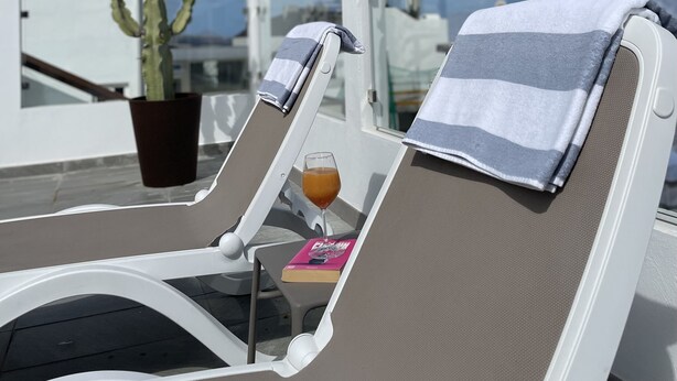 A sun lounger with a towel, a glass of drink, and a book on a balcony with a cactus and a clear sky. - Hotel Idafe (Las Palmas de Gran Canaria)