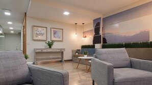 A modern living room with grey sofas, a coffee table, and a dining area with a small table and chairs. There are framed artworks on the walls and a large window with a view of the outdoors. - Hotel Idafe (Las Palmas de Gran Canaria)