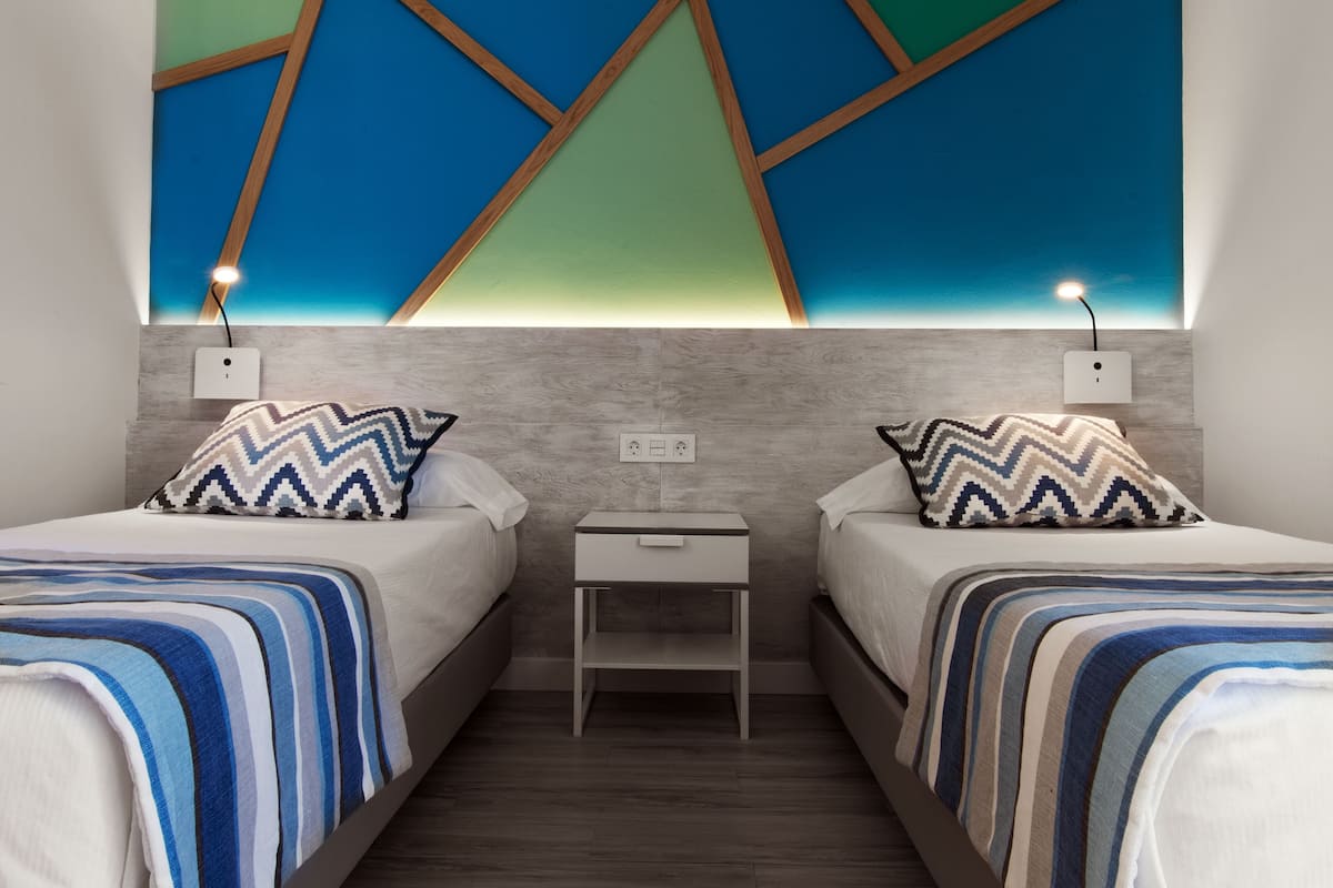 A modern bedroom with two beds, a nightstand, and a wall-mounted light fixture.
