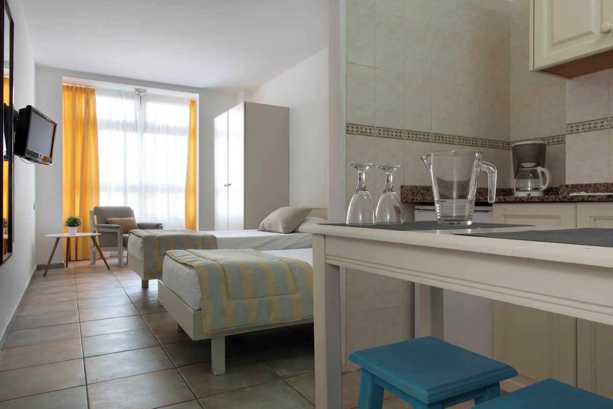 A hotel room with a bed, a chair, a small table, a kitchen area with a coffee maker, and a TV mounted on the wall.