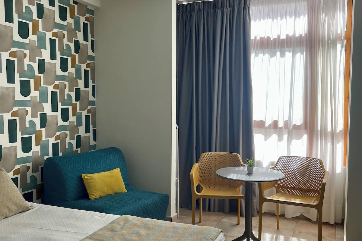 A hotel room with a bed, a blue sofa, a yellow chair, a small table, and a window with curtains.