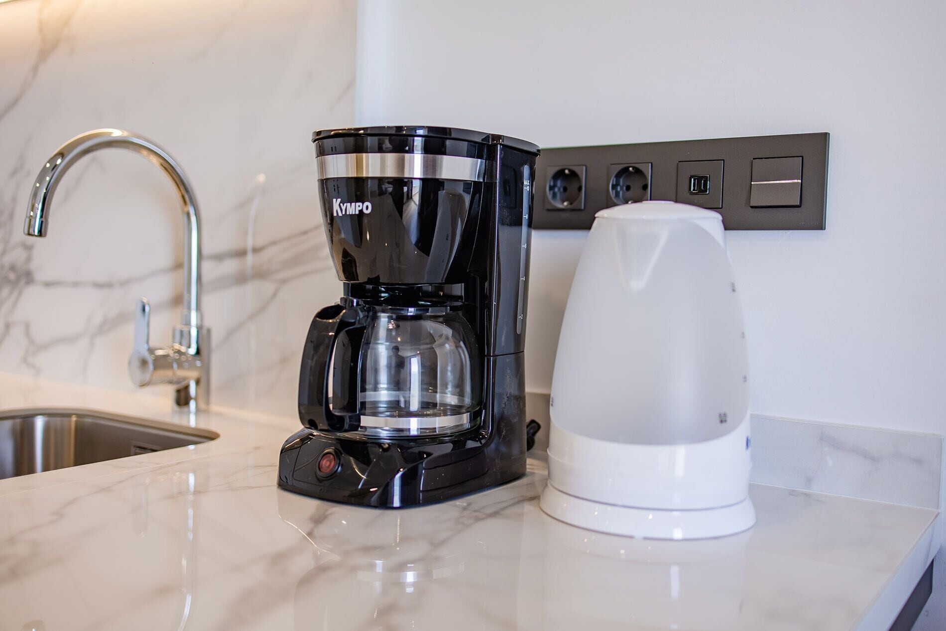 A modern kitchen with a black coffee maker, a white electric kettle, and a marble countertop.