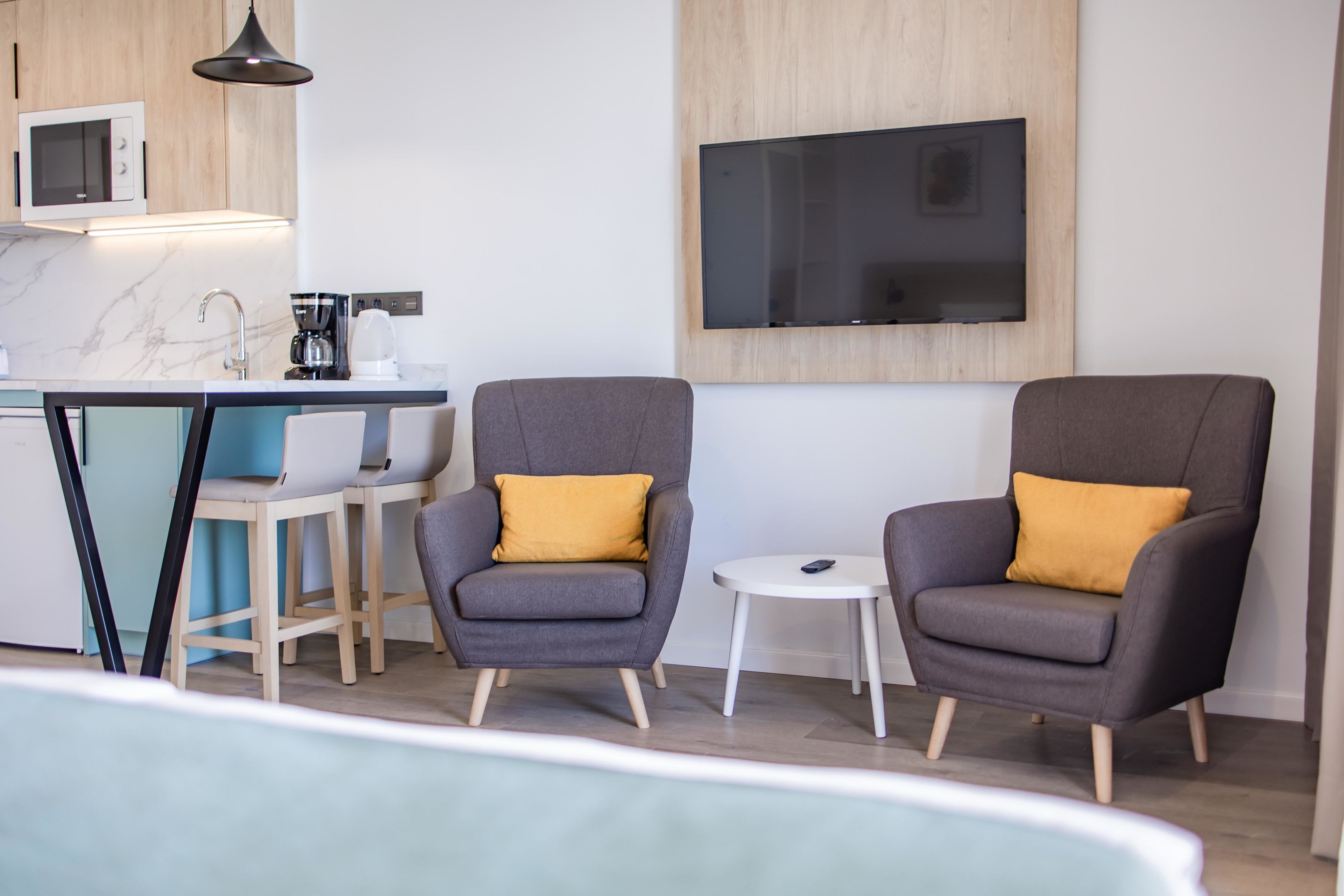 A modern kitchen with a microwave, coffee maker, and a large flat-screen TV mounted on the wall. There are two grey armchairs with yellow cushions and a small round table.