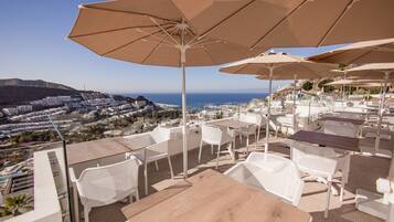 Una terraza con sillas y mesas blancas, grandes sombrillas y vistas a la costa y edificios.