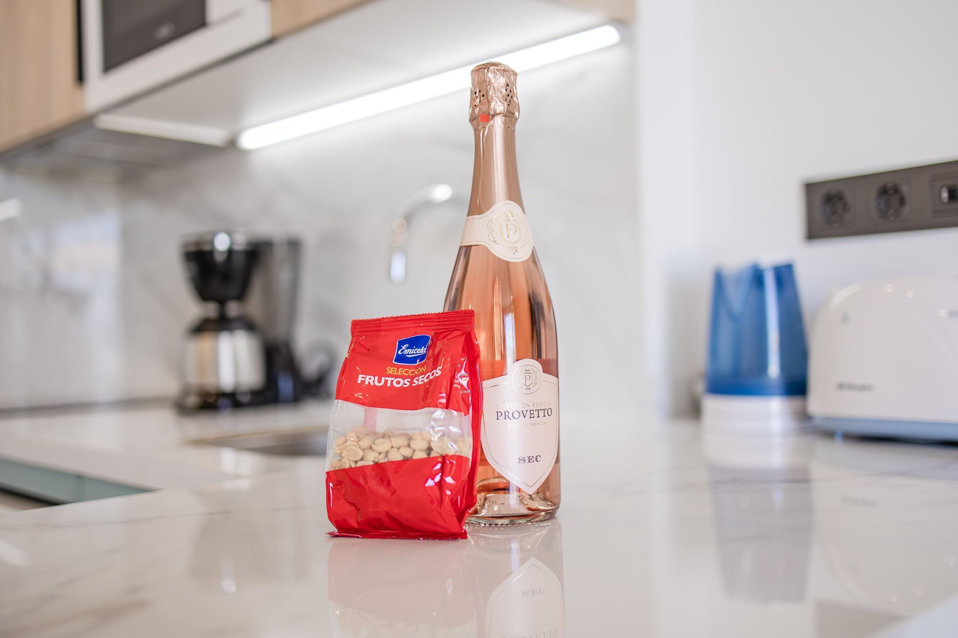 A kitchen counter with a bottle of Provettto Sec sparkling wine, a red bag of Emicola Selecta fruits, and a coffee maker.