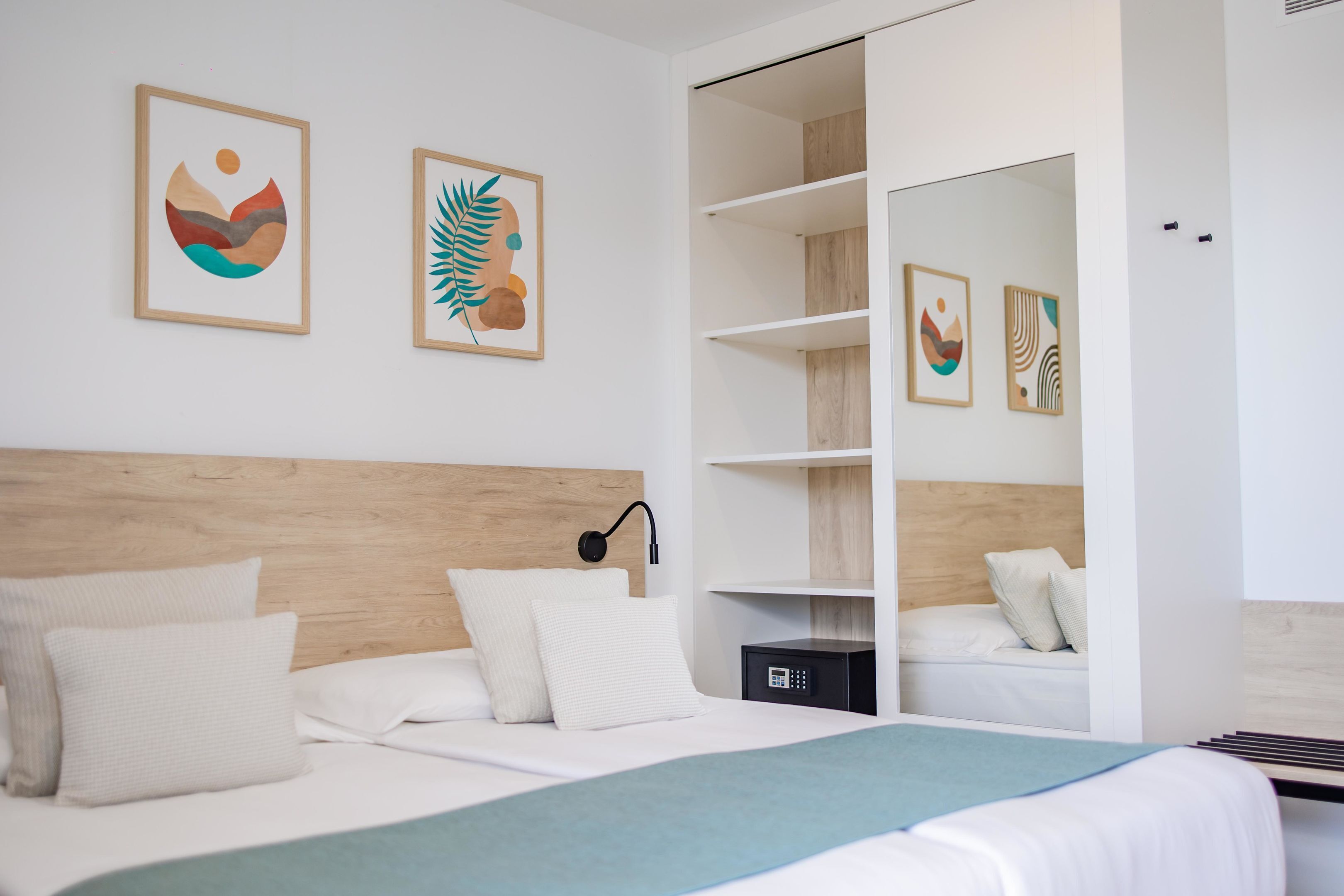 Una habitación de hotel moderna con cabecera de madera, cama con ropa blanca, manta azul y armario empotrado con estantes.