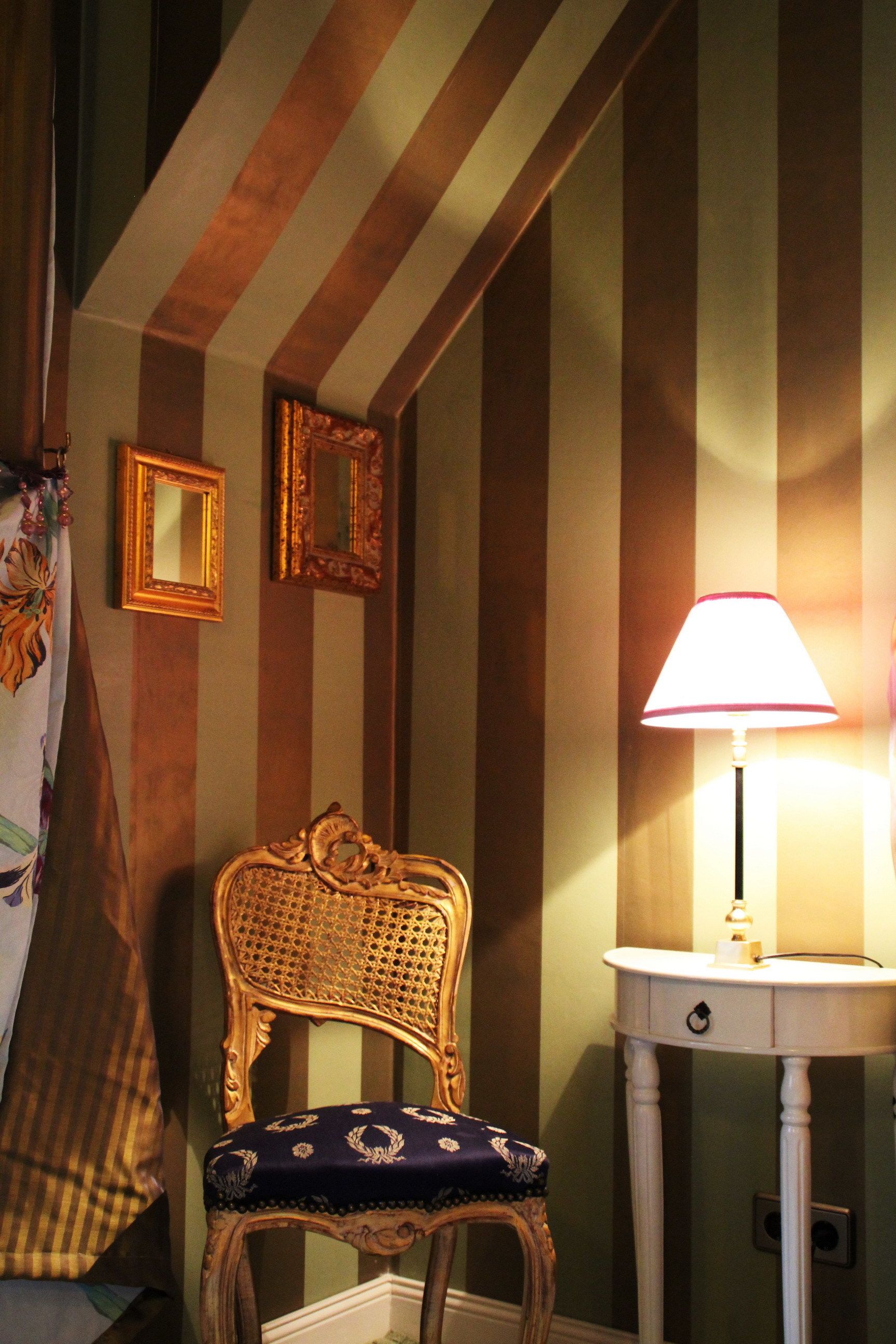 Una habitación con papel tapiz a rayas, una silla decorativa, una mesita de noche con una lámpara y dos espejos enmarcados en la pared.
