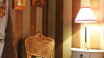 Una habitación con papel tapiz a rayas, una silla decorativa, una mesita de noche con una lámpara y dos espejos enmarcados en la pared.
