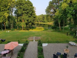 Un jardín bien cuidado con un estanque central, rodeado de abundante vegetación y árboles. Hay zonas de estar al aire libre con mesas y sillas.