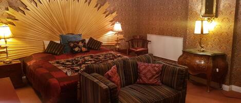 A bedroom with a large bed, two bedside tables with lamps, a chair, a small table, and a mirror on the wall.