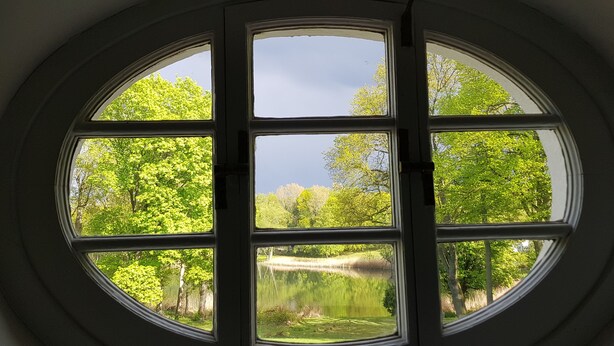 A circular window with six panes, offering a view of a pond surrounded by lush green trees. - Villa am Stechlin (Stechlin)
