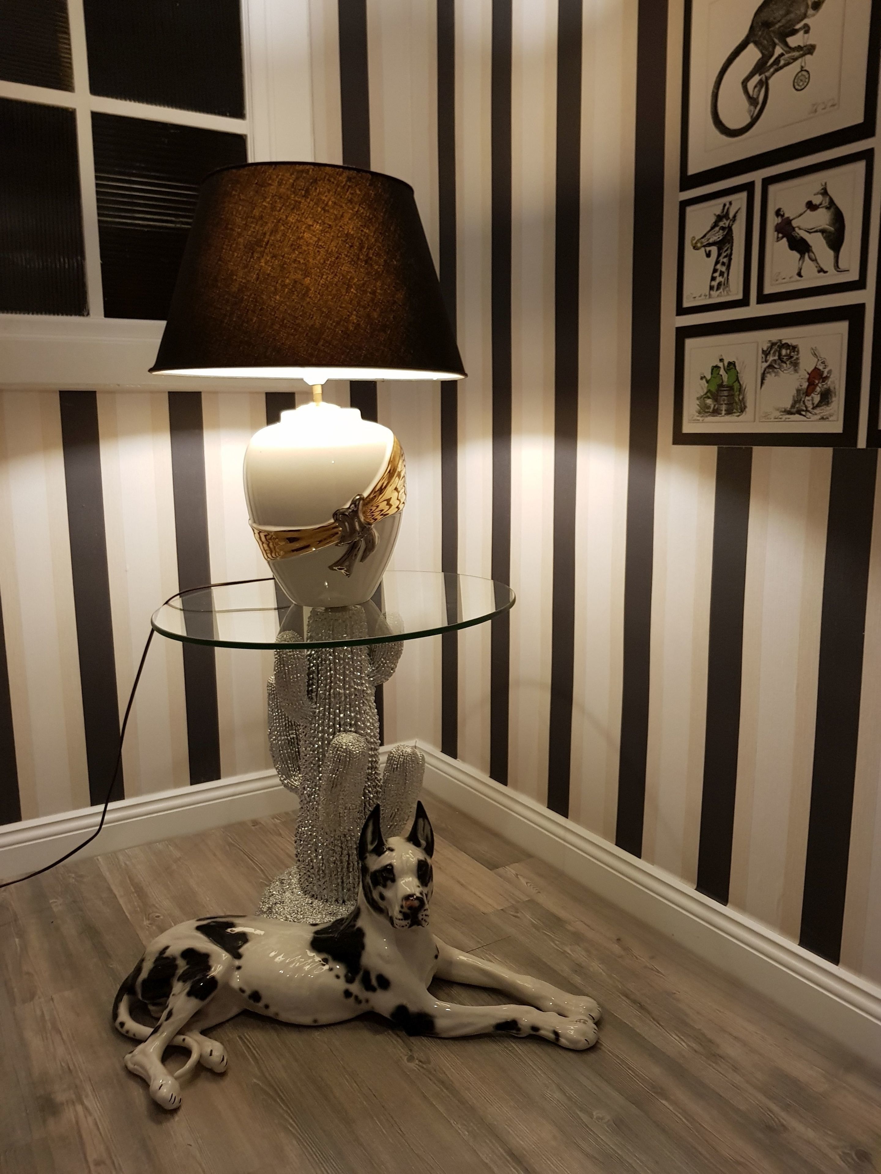 A lamp with a gold base and black lampshade on a glass table, a black and white dog lying on the floor, and a wall with striped wallpaper and framed pictures.