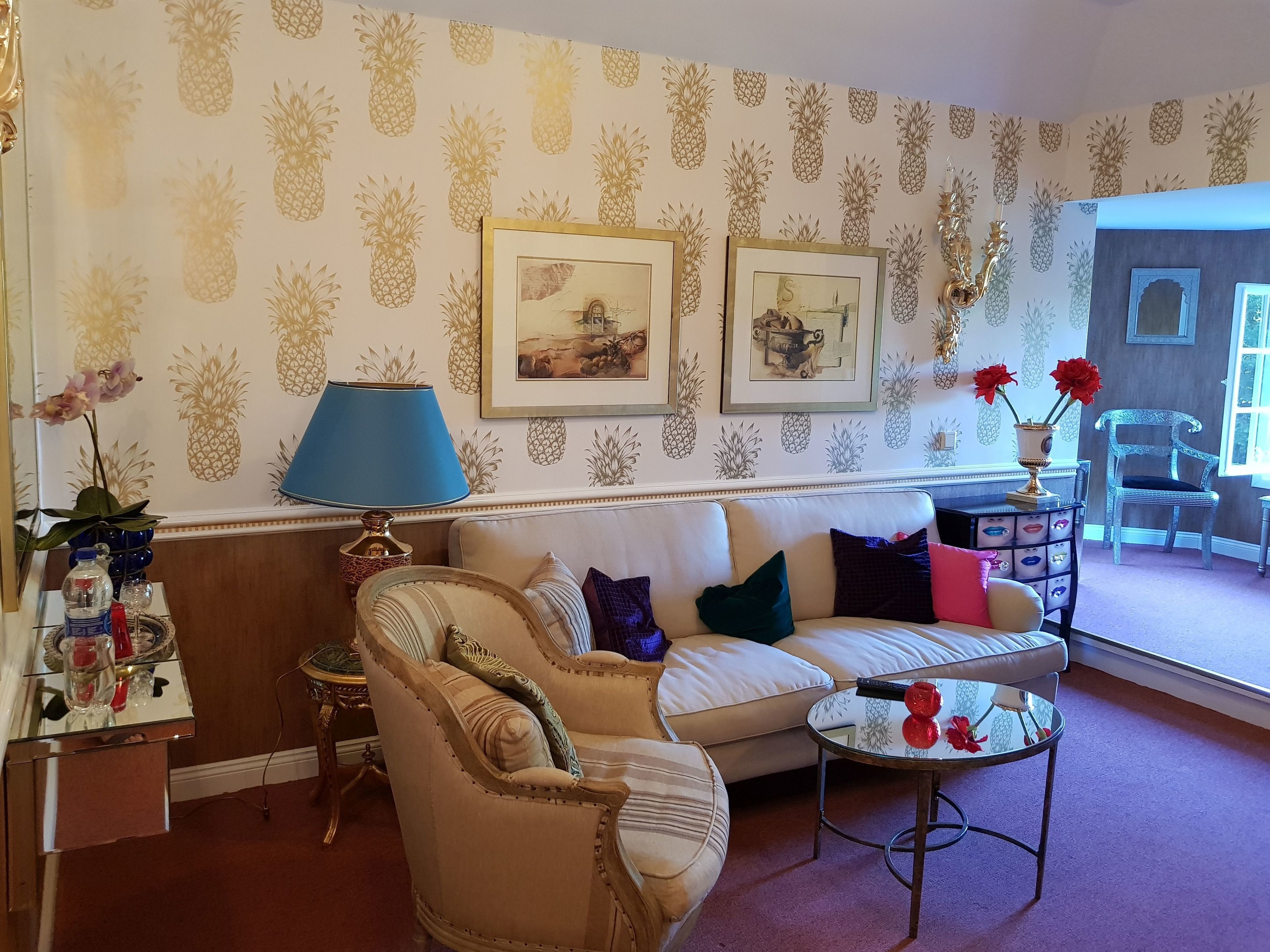 A living room with a patterned wallpaper, a sofa, a chair, a small table with decorative items, and framed pictures on the wall.