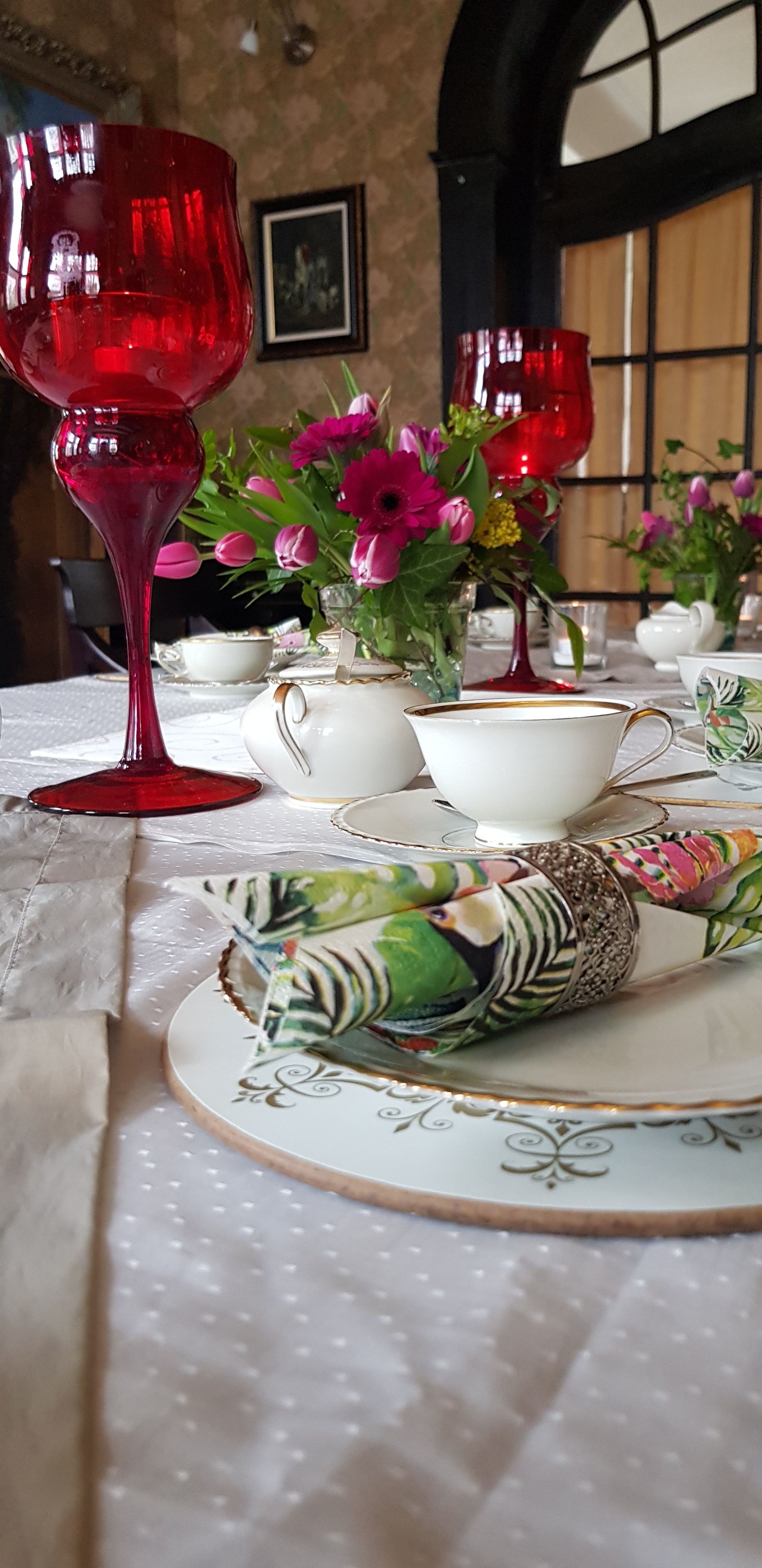 A table set with a red glass, white teapot, and a colorful bird-shaped napkin holder.