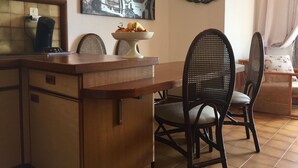 Un coin repas avec une table en bois, deux chaises en osier, un saladier rempli de fruits et un tableau représentant un vélo accroché au mur.