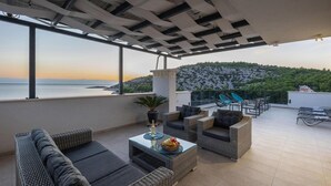 Um terraço com mobiliário de vime, uma mesa de vidro com uma cesta de frutas, e vista para o mar e colinas.