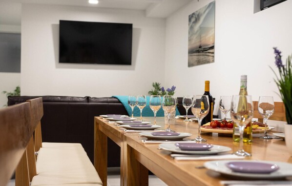 Un coin repas avec une longue table en bois dressée pour le repas, un canapé en cuir noir et un téléviseur à écran plat fixé au mur.