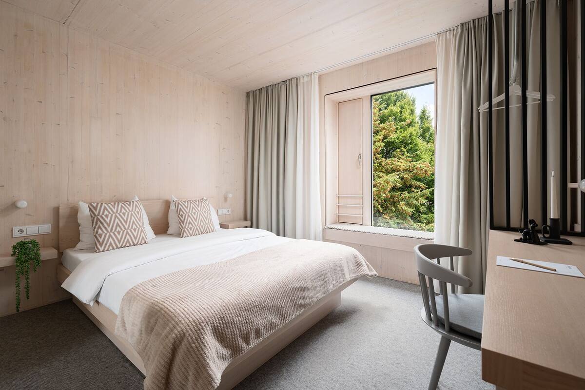 A bedroom with a bed, a desk, a chair, a window with curtains, and a view of trees.