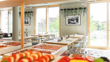A restaurant with multiple tables, a variety of food displayed on the counter, and large windows with a view of greenery outside.