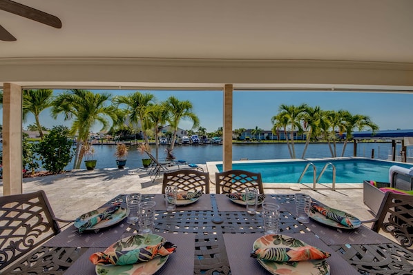 Une table avec des chaises placée au bord de la piscine, avec vue sur la piscine et des palmiers.