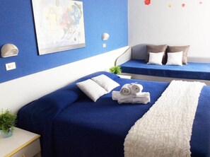 A hotel room with two beds, each with blue bedding and white pillows. There is a framed picture on the wall, a small plant on the nightstand, and a white fluffy throw on one bed. - Hotel Augustus Milano Marittima (Cervia)