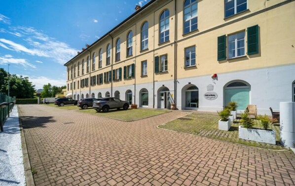 A large, multi-story building with a yellow facade and green shutters, featuring a paved driveway and parked cars. - Bluelake Inn - Ostello (Olginate)